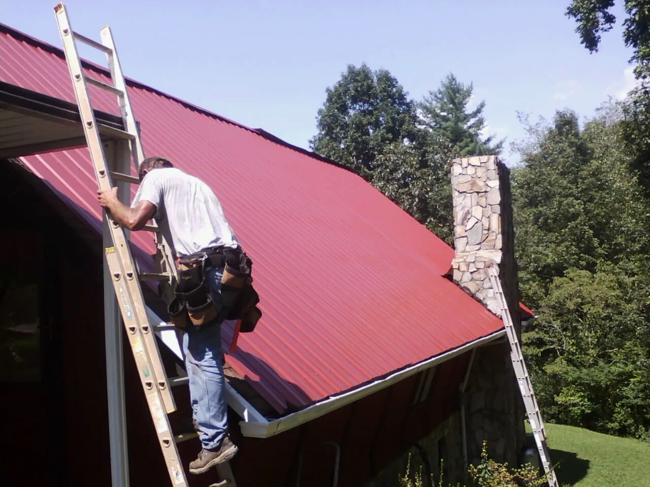 Roofer on a ladder beside a red metal roof and stone chimney — Soffit & Fascia Repair by Roofing Brothers in Verplanck